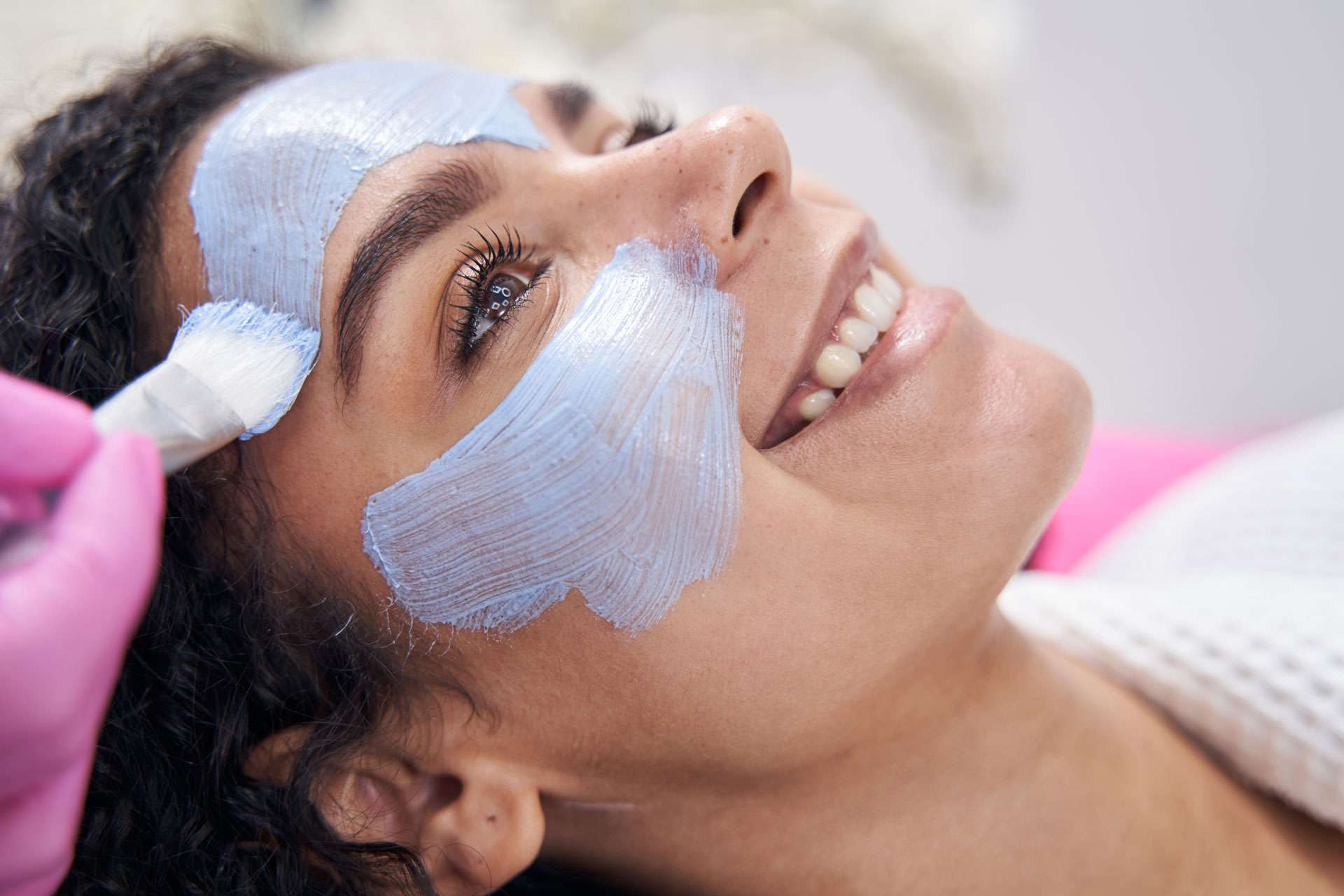 Person reclining during a facial treatment as a brush applies a white facial mask to the forehead and cheek